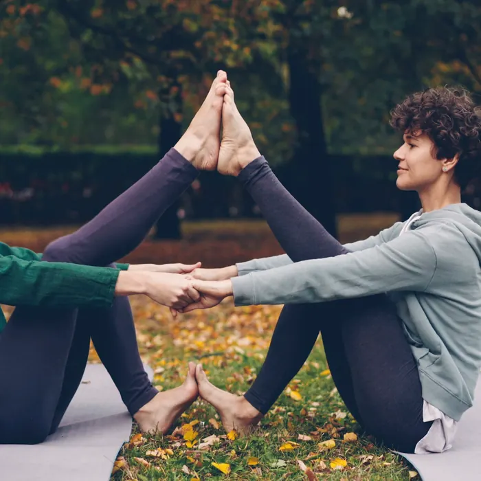 Two friends sit facing each other in autumn leaves and stretch, holding hands.