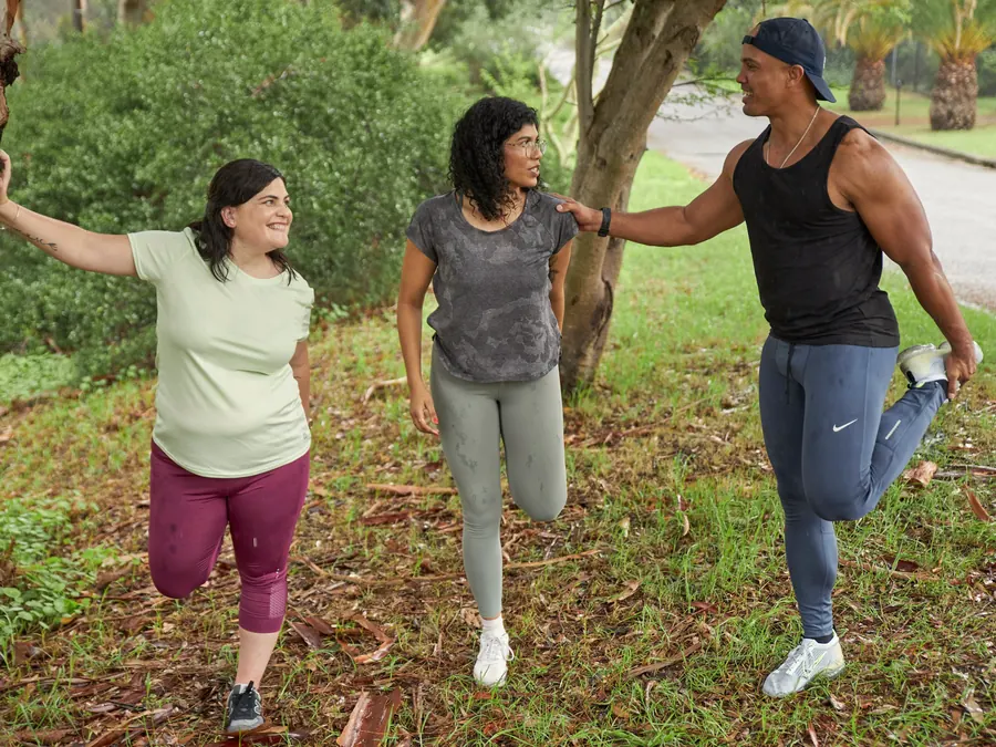 Three friends laugh while they stretch their legs in a park before a run.