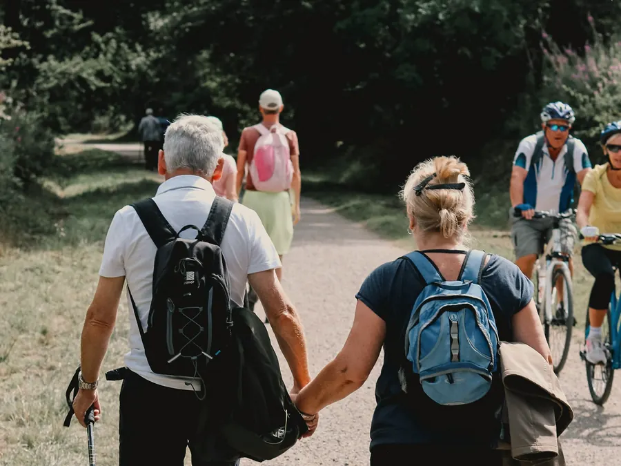 A couple with backpacks and walking poles hike along a path, hand in hand.